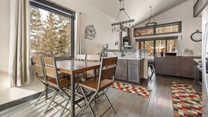 A modern kitchen with a dining area, wooden chairs, and a large window with a view of trees.