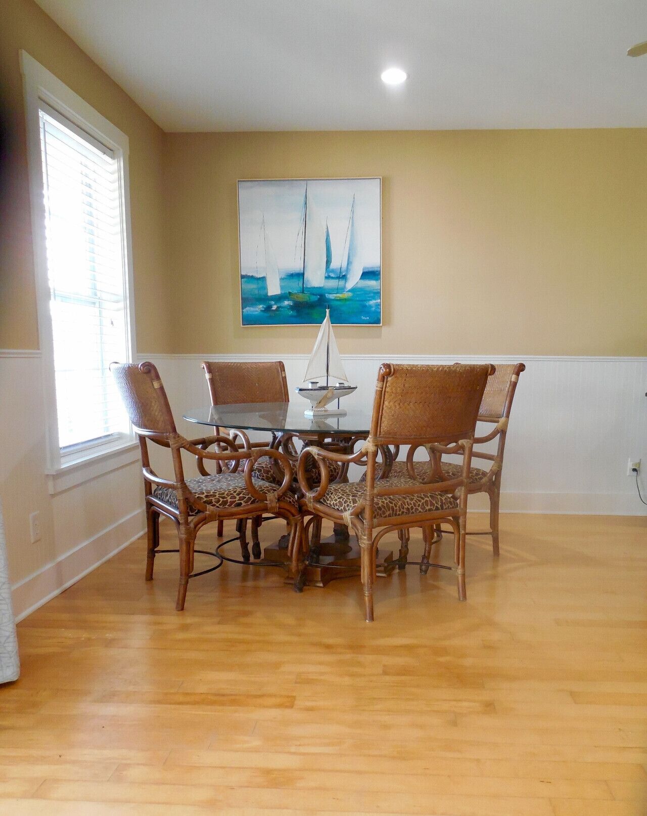 A dining area with a glass table surrounded by four wicker chairs with leopard print cushions. A sailboat model is on the table, and a painting of sailboats is on the wall.