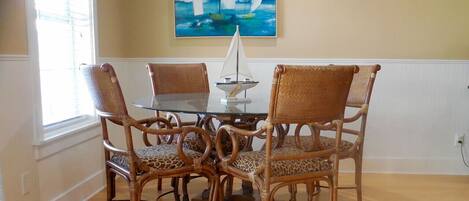 A dining area with a glass table surrounded by four wicker chairs with leopard print cushions. A sailboat model is on the table, and a painting of sailboats is on the wall.