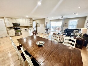 A spacious living area with a large wooden dining table, white chairs, a kitchen with white cabinets, and a balcony with a view.