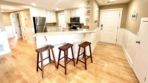 A modern kitchen with white cabinetry, a stainless steel refrigerator, a microwave, and a large island with bar stools. - Tybee Retreat Unit 3 (Tybee Island)