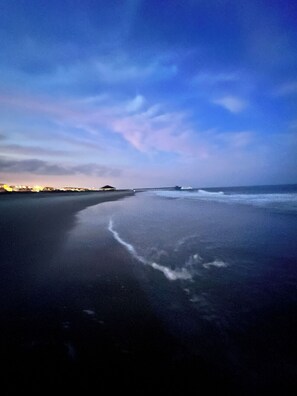 A beach with a calm sea, a pier in the distance, and a clear sky.