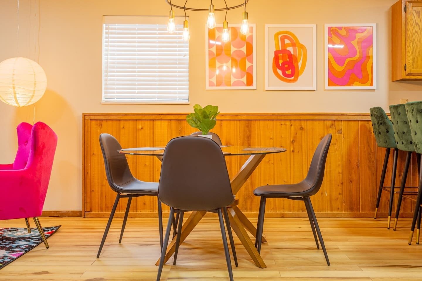 A dining area with a wooden wall, a round table, and four chairs. There are two windows with blinds, a hanging light fixture, and two framed artworks on the wall.