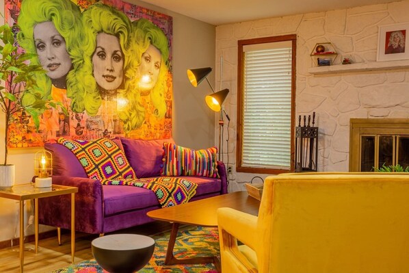 Ein lebhaftes Wohnzimmer mit einem violetten Sofa, bunten Kissen, einem gelben Sessel, einem schwarzen Couchtisch und einem großen abstrakten Gemälde an der Wand.
