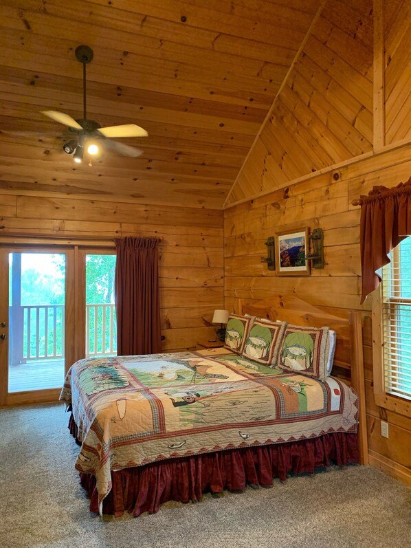 A wooden cabin bedroom with a large bed, a ceiling fan, and a view of the outdoors.