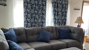Un canapé modulable avec des coussins bleus, un pouf assorti et une fenêtre ornée de rideaux à motifs floraux.