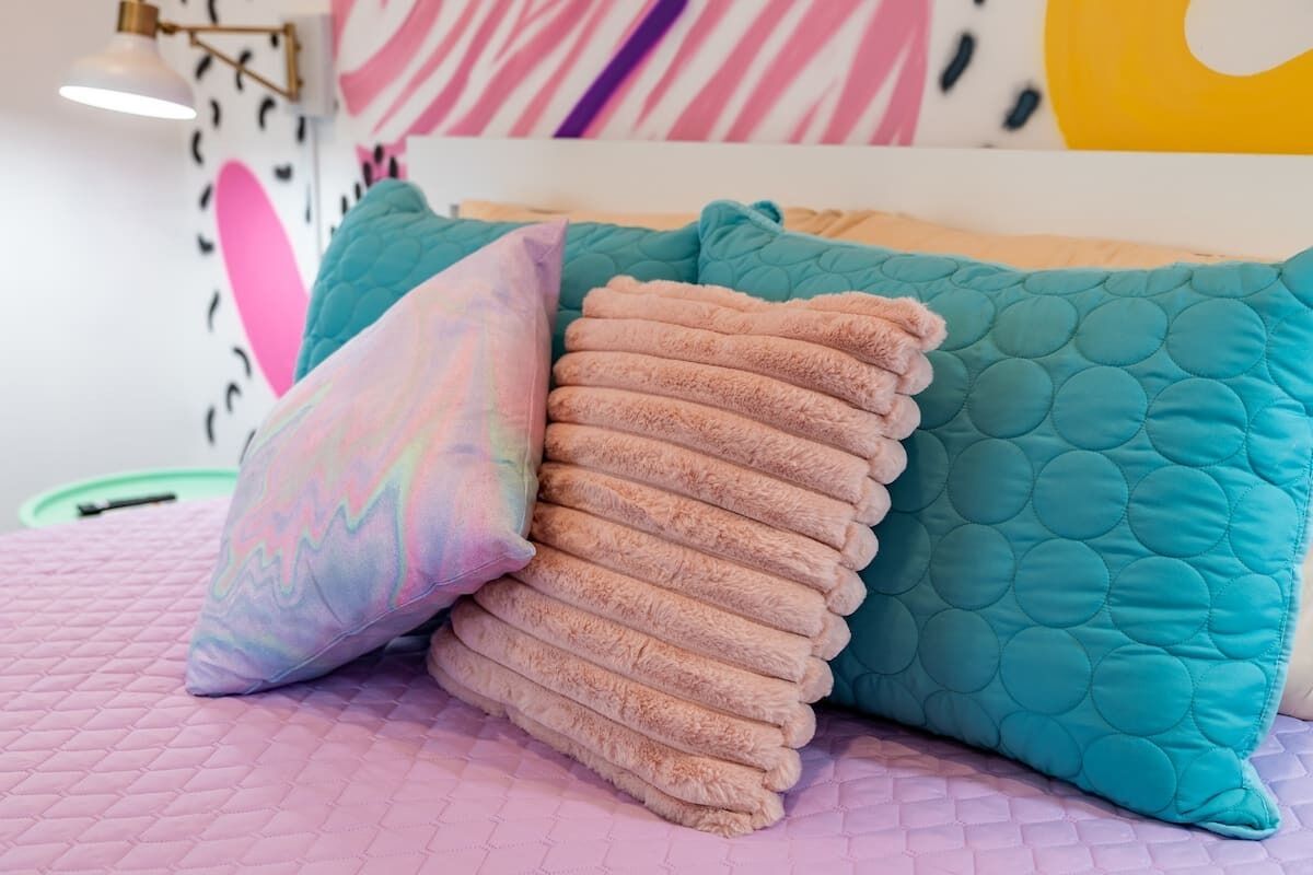 A bed with teal and pink pillows, a pink quilted bedspread, and a wall with abstract art.