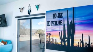 A room with a blue sofa, a sliding glass door leading to an outdoor area, and a poster with cacti and a sunset. - ❤Hot Tub + Close to Downtown JT ❤ Fully Fenced + Firepit Mini❤ (Joshua Tree)