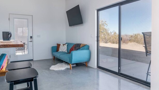 A modern living room with a teal sofa, a flat-screen TV, and a large sliding glass door leading to an outdoor area. - ❤Hot Tub + Close to Downtown JT ❤ Fully Fenced + Firepit Mini❤ (Joshua Tree)