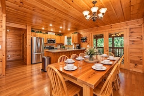 A wooden cabin interior with a dining table set for four, kitchen appliances, and a view through the windows. - ❤Family Friendly❤3 Min to Parkway❤Gameroom, Hot Tub, Theater❤ (Pigeon Forge)