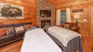 Un interior de cabaña acogedor con una chimenea de piedra, un sofá con cojines estampados, un televisor montado en la pared y una cama con una colcha.