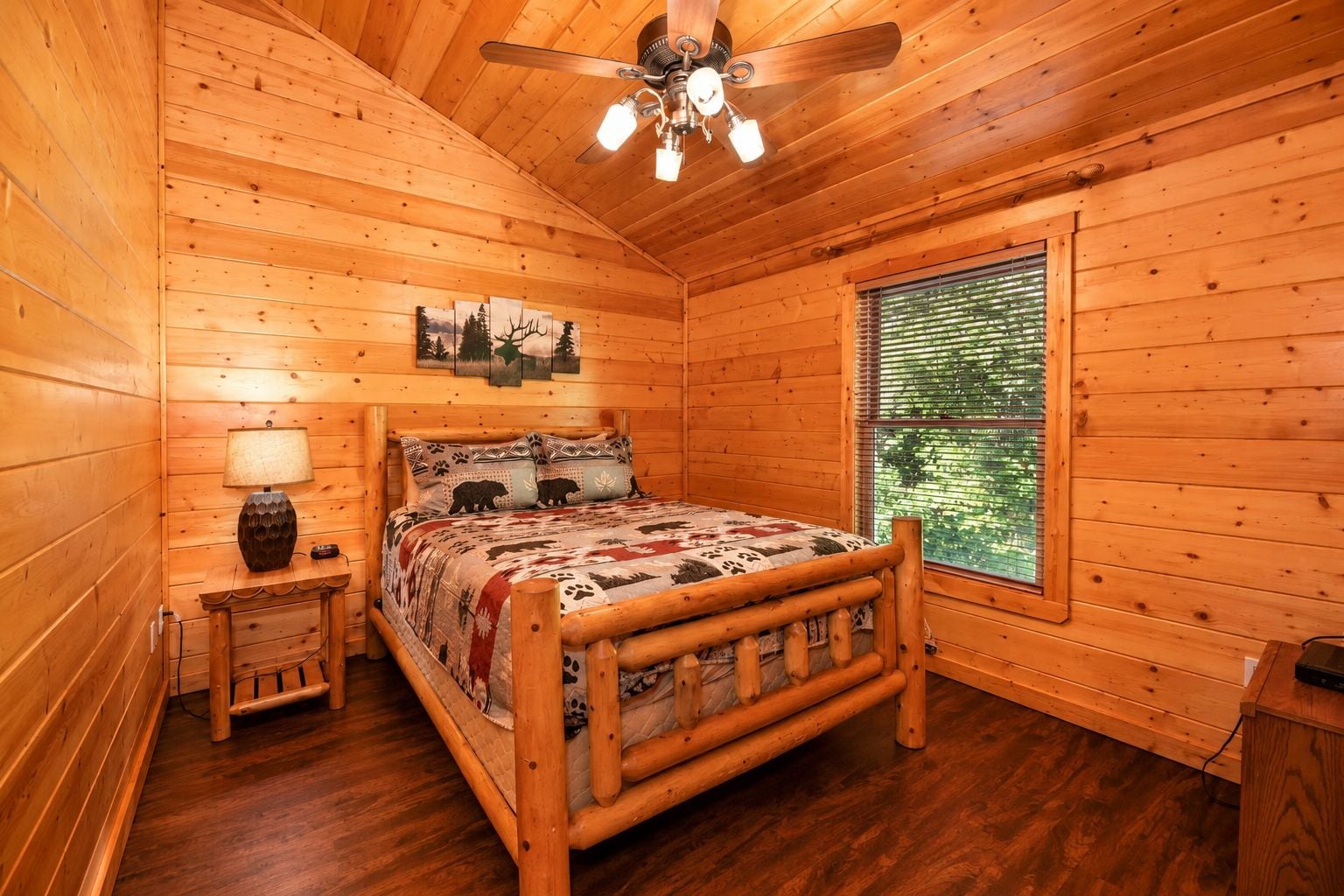 A wooden cabin bedroom with a bed, a nightstand, a lamp, a ceiling fan, and a window with blinds.