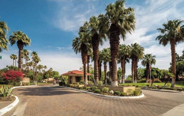 A paved entrance with palm trees and a small building with a red roof.