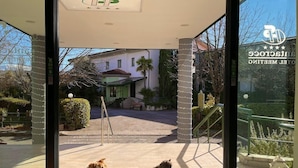 L’entrée d’un hôtel, avec une porte en verre, un tapis vert portant le nom de l’hôtel, et deux chiens assis sur le tapis.