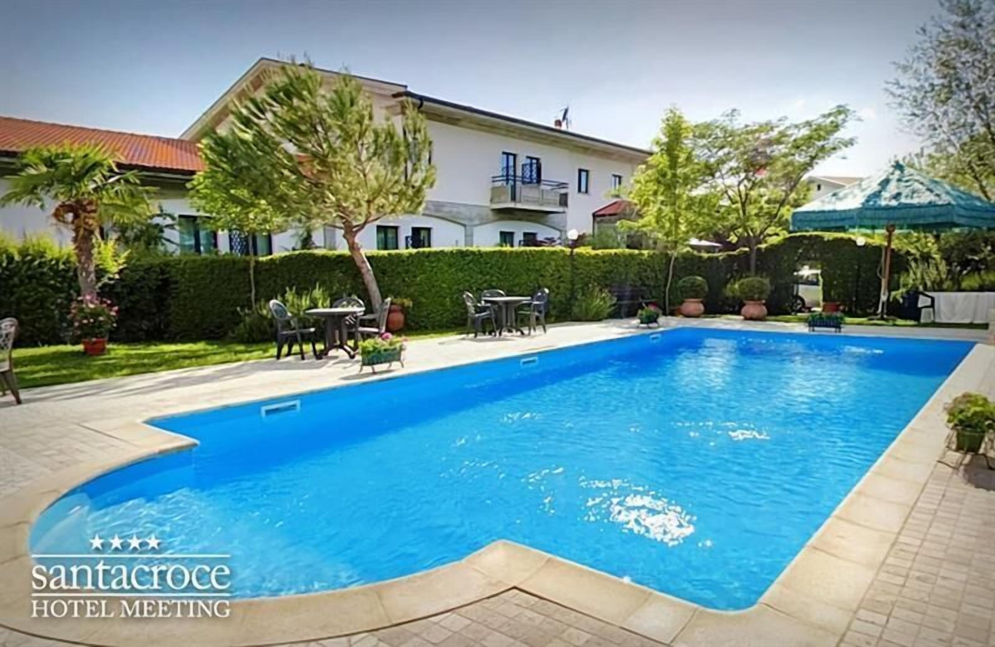 A swimming pool with a tiled deck, surrounded by a well-maintained garden and a building with balconies.