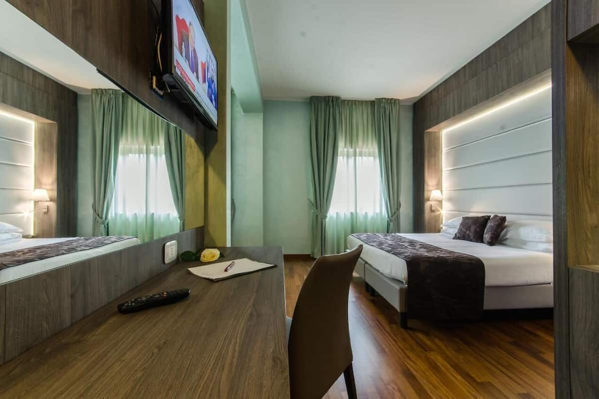 Habitación de hotel con cama, escritorio con silla, televisor montado en la pared y cortinas en las ventanas.
