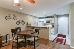 A compact kitchen with white cabinets, a microwave, and a refrigerator. There is a dining area with a wooden table and chairs.