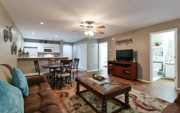 A living room with a brown sofa, a glass coffee table, a dining area with a table and chairs, a kitchen with white cabinets, a ceiling fan, and a TV on a wooden cabinet. - Shelter in Faith- Amazing Golf Views! (Branson)