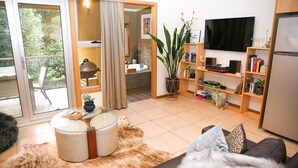 Ein modernes Wohnzimmer mit einer Couch, einem Couchtisch und einem Fernseher. Ein Bücherregal mit Büchern und Dekorationselementen ist zu sehen. Durch eine offene Tür ist ein Badezimmer erkennbar.