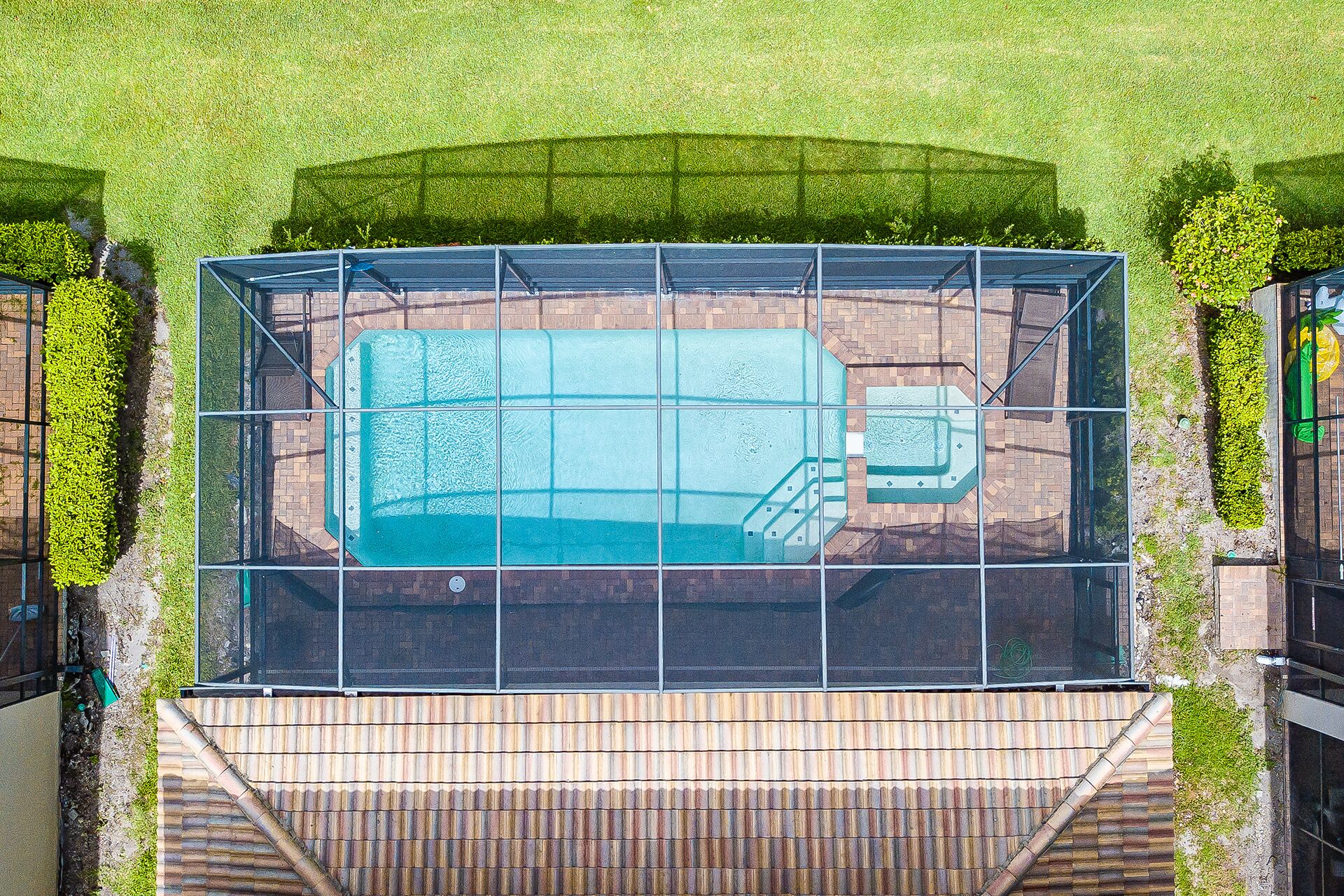 Une vue aérienne d’une piscine entourée d’une terrasse, délimitée par une clôture en métal, et située à côté d’un bâtiment à la façade en briques.