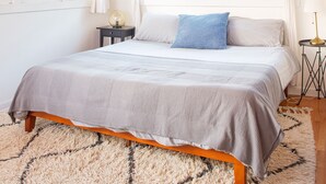 Une chambre avec un plafond en bois, un lit recouvert d’une couette grise et blanche, un tapis moelleux et deux fenêtres avec des rideaux.