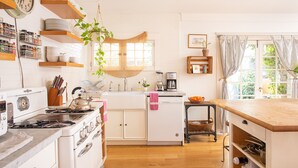 Une cuisine avec des meubles blancs, une cuisinière et une machine à café. On y trouve des étagères remplies de vaisselle et une plante suspendue au plafond.