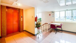 Um saguão de hotel com elevador vermelho, balcão de recepção, planta em vaso e janela com vista para a cidade.