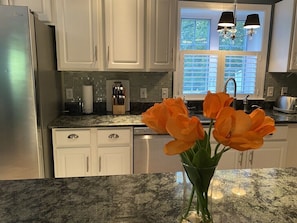 Eine Küche mit weißen Schränken, einem Edelstahl-Kühlschrank, einer Arbeitsplatte aus Granit und einer Vase mit orangefarbenen Blumen.
