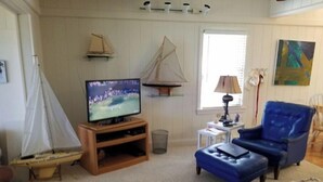 A living room with a television, a blue armchair, a wooden entertainment unit, a small table with a lamp, and a sailboat model. - Pappy's Pool Lake: A  1650 sqft with Pier,  Boat Ramp, Screened porch,Lakeside. (Lake Waccamaw)