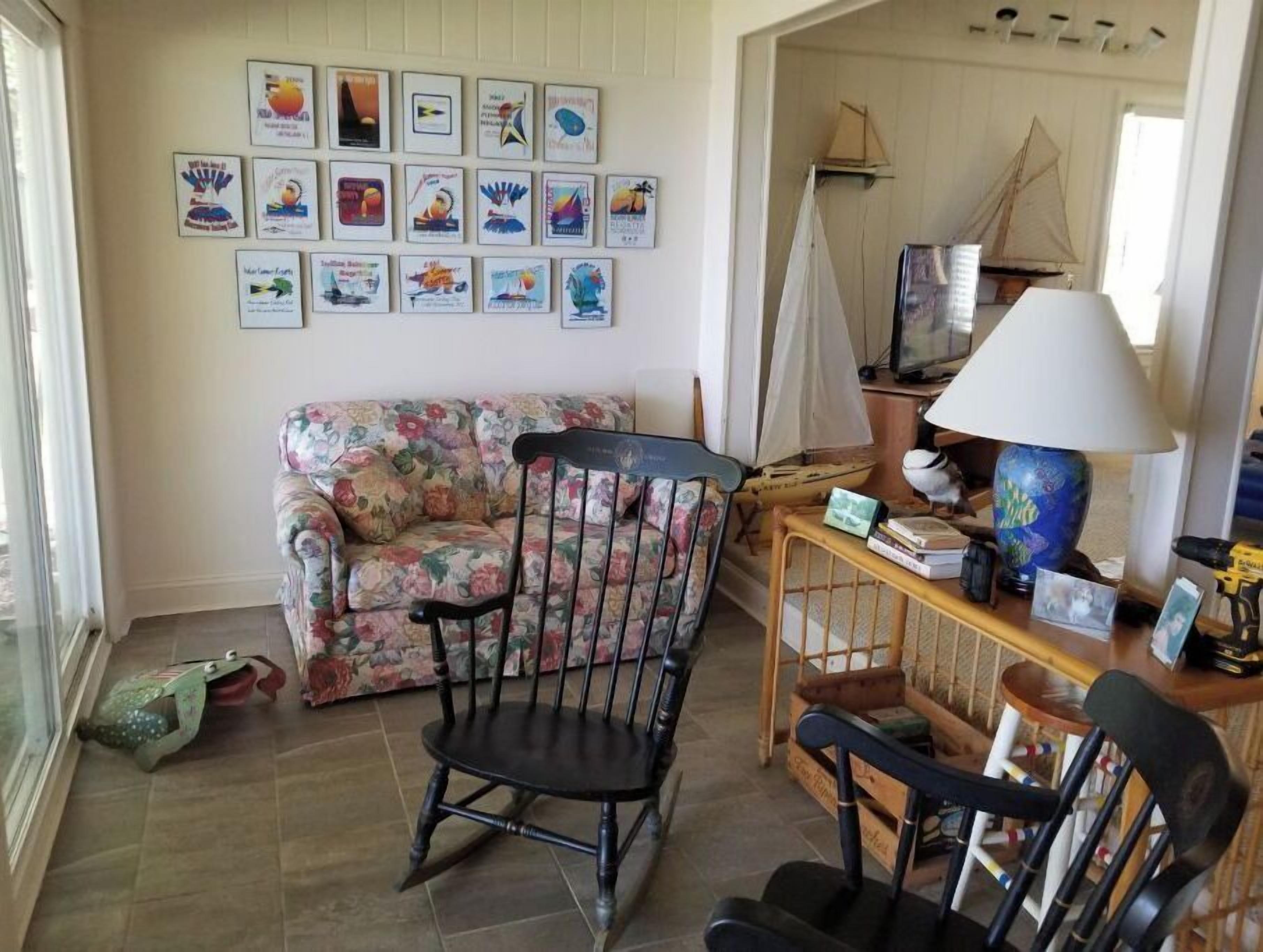 Een woonkamer met een bloemenbank, een houten schommelstoel, een kleine tafel met een lamp, en een wand versierd met ingelijste kunstwerken.