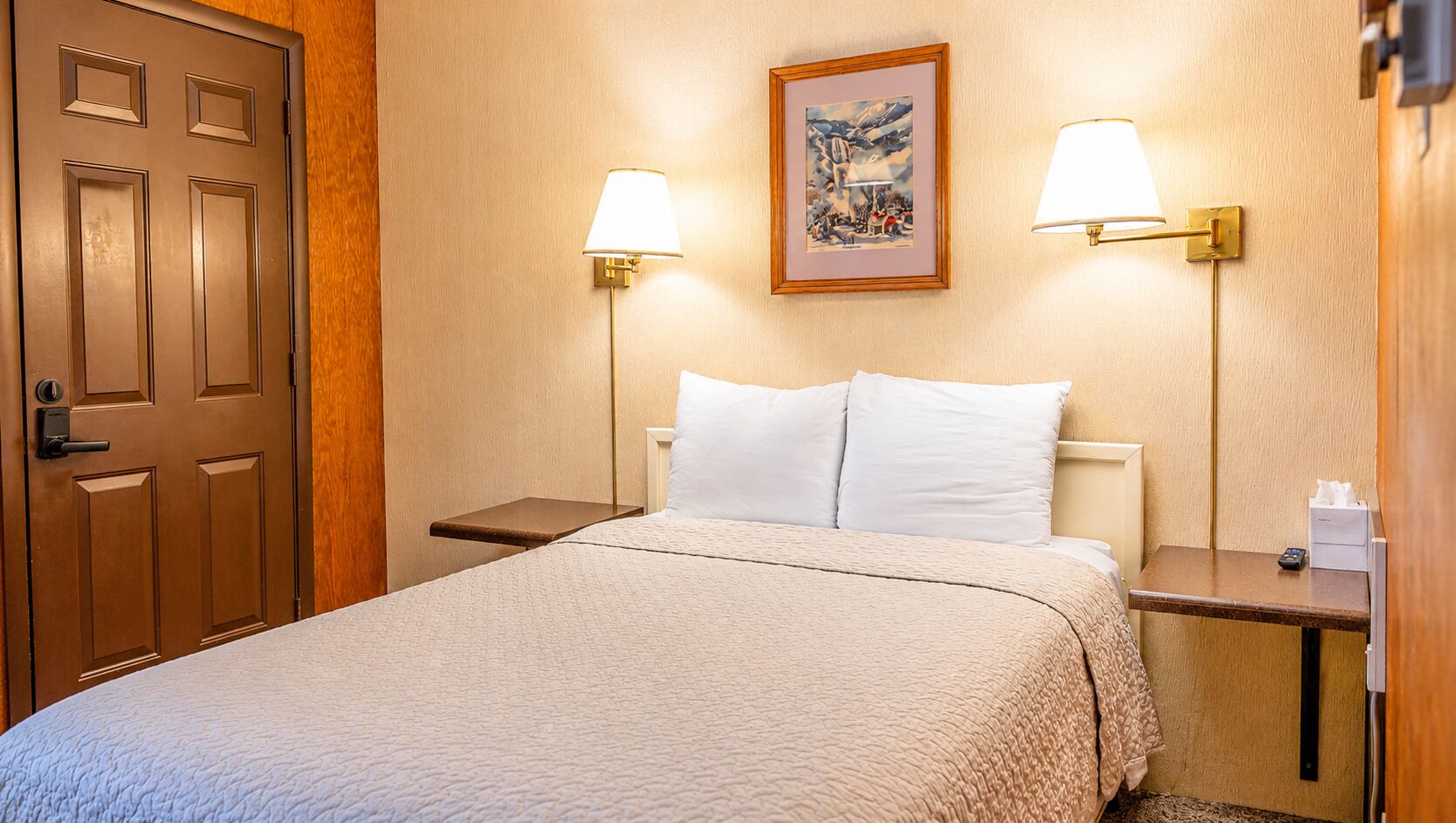 Habitació d'hotel amb un llit, dues taules de nit, una porta i dues làmpades penjades a la paret.