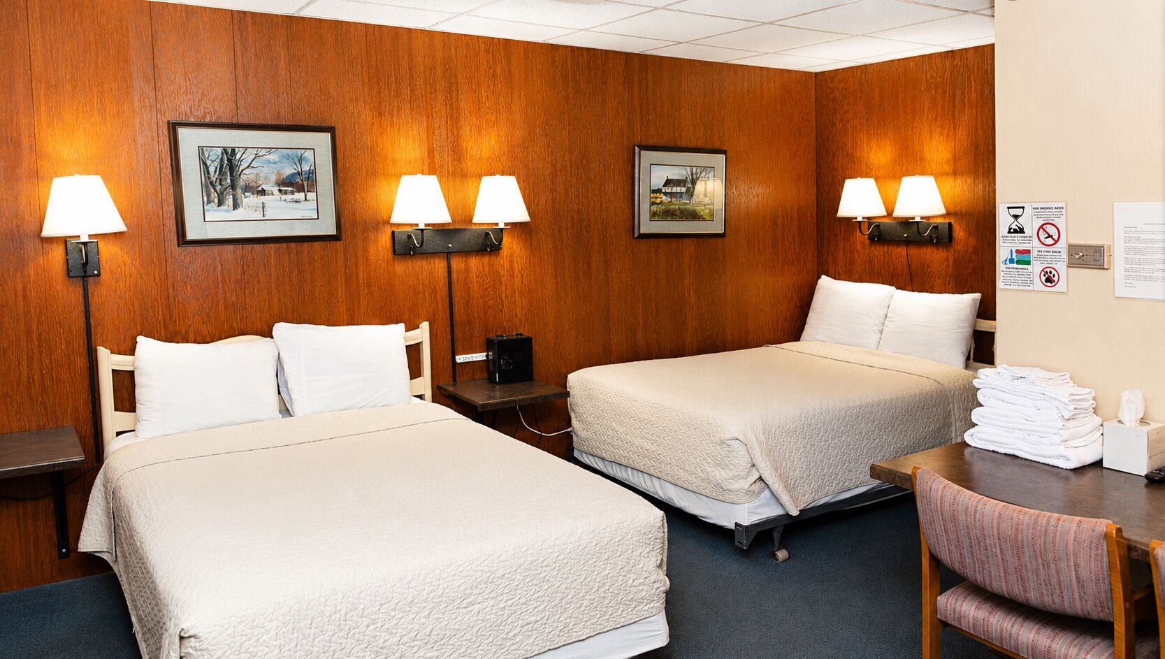 A hotel room with two beds, a desk, and a chair. There are wall-mounted lamps and framed pictures.