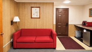 Una habitación con un sofá rojo, paredes de paneles de madera, una puerta, un cuadro enmarcado, una lámpara, un microondas y un cubo de basura.