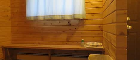 Una cabaña de madera con un banco, una ventana con cortina, una mesita con una botella y un cubo de basura.