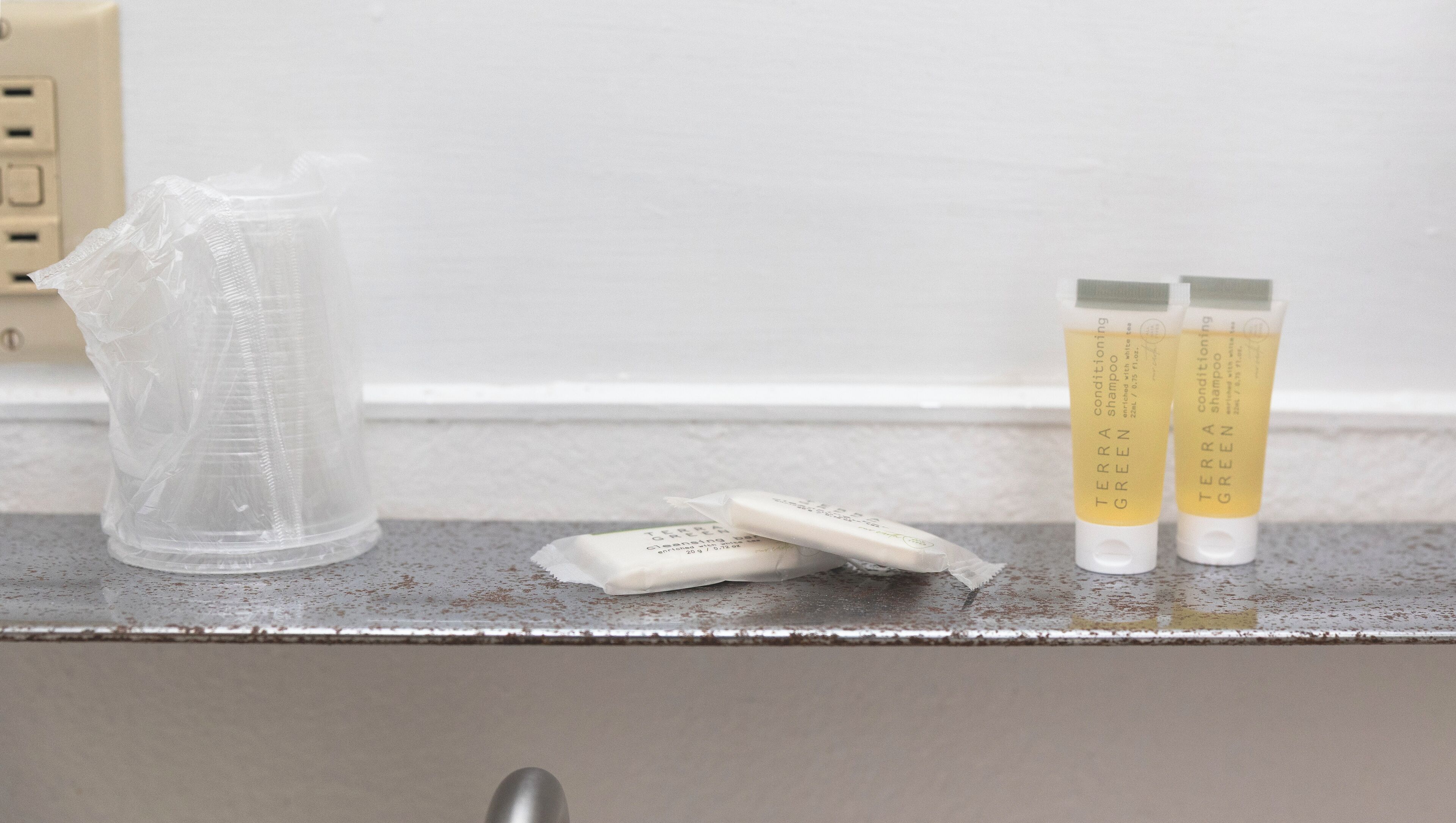 Artigos de higiene pessoal em uma bancada de banheiro, incluindo um recipiente de plástico transparente, dois tubos de shampoo e uma barra de sabão.