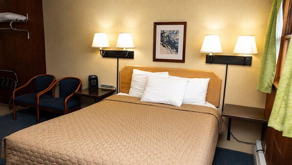 A hotel room with a bed, two chairs, a bedside table, a lamp, and a painting on the wall.