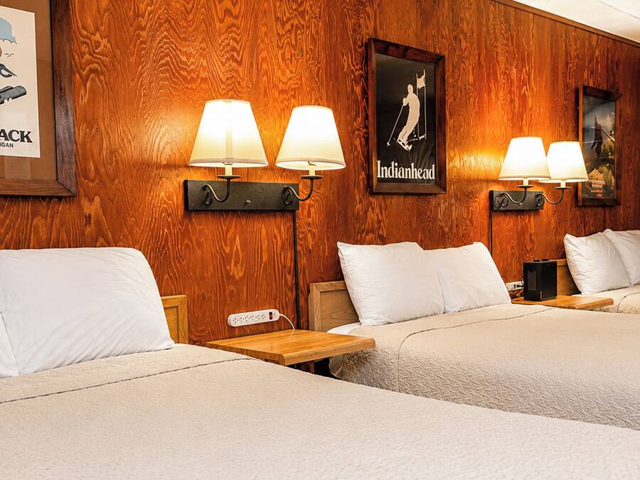 A hotel room with wooden paneling, three beds, wall-mounted lamps, and two posters.