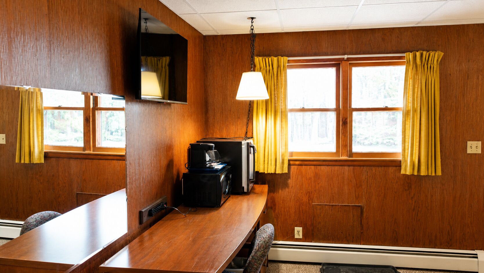 Een kamer met een houten wand, een aan de muur bevestigde televisie, een tafel met een stoel en een raam met gordijnen.