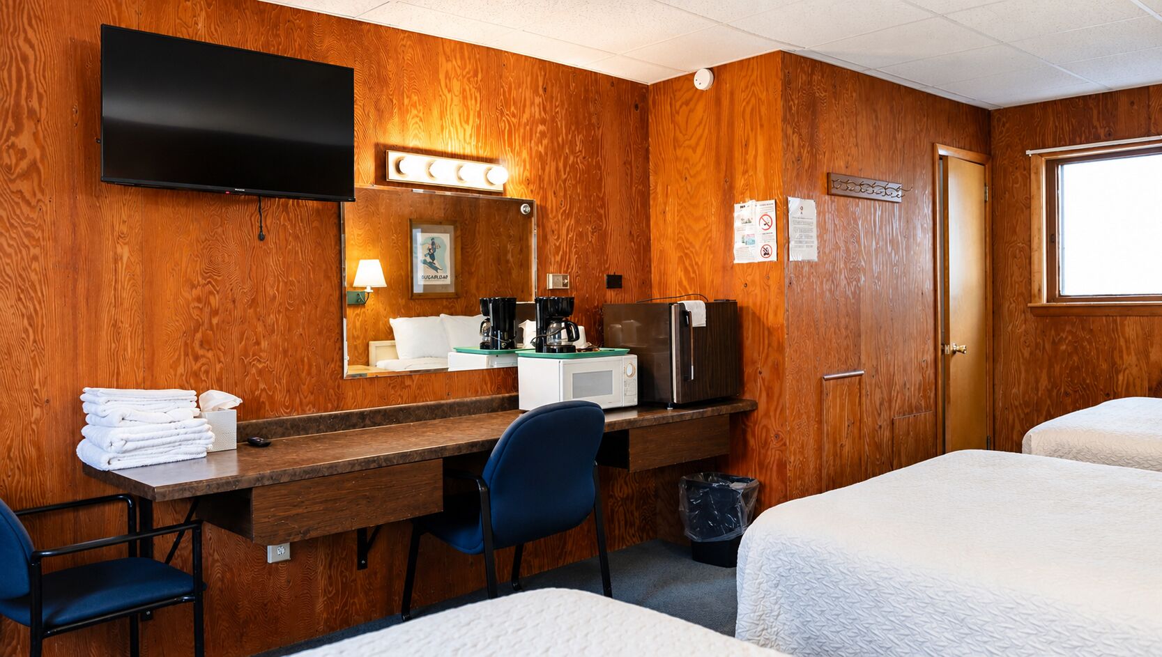 A hotel room with a wooden wall, a flat-screen TV, a desk with a chair, a microwave, a refrigerator, and two beds.