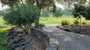 A paved pathway leading to a golf course with a stone wall on one side and a variety of trees and shrubs. - Modern Condo in Gated Silverado Resort Residential Community (Napa)