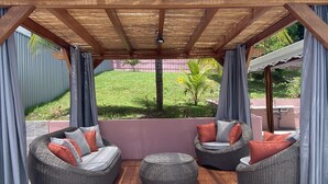 Uma área de estar coberta ao ar livre, com móveis de vime e vista para um jardim.