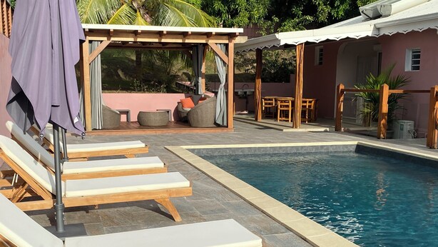 Uma área de piscina com espreguiçadeiras, um pergolado e uma pequena mesa com cadeiras.