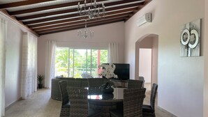 Uma sala de jantar com mobiliário de vime, um candelabro e uma grande janela com cortinas.