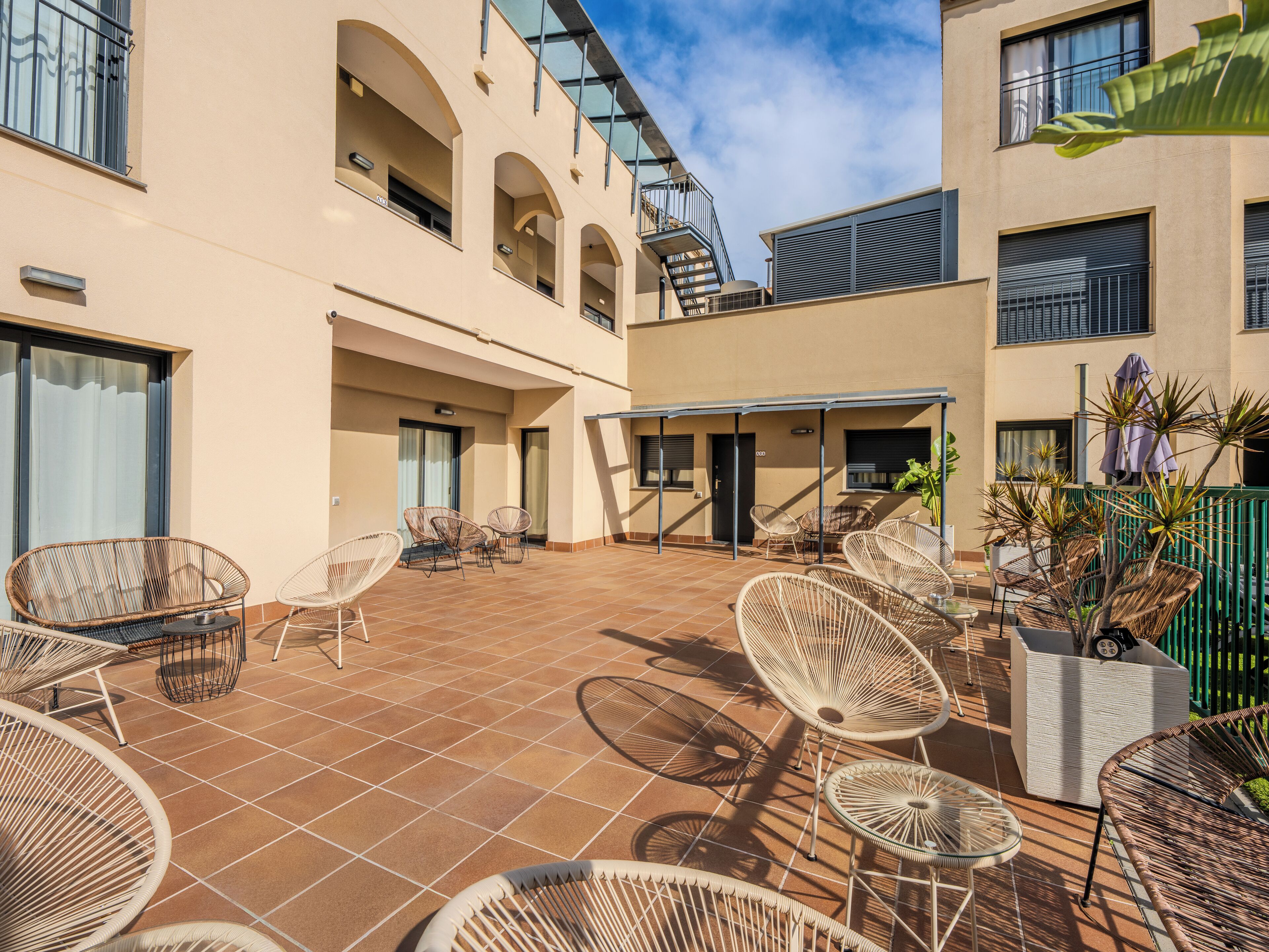 Un patio con sillas de mimbre, un espacio de descanso techado y un edificio con balcones.