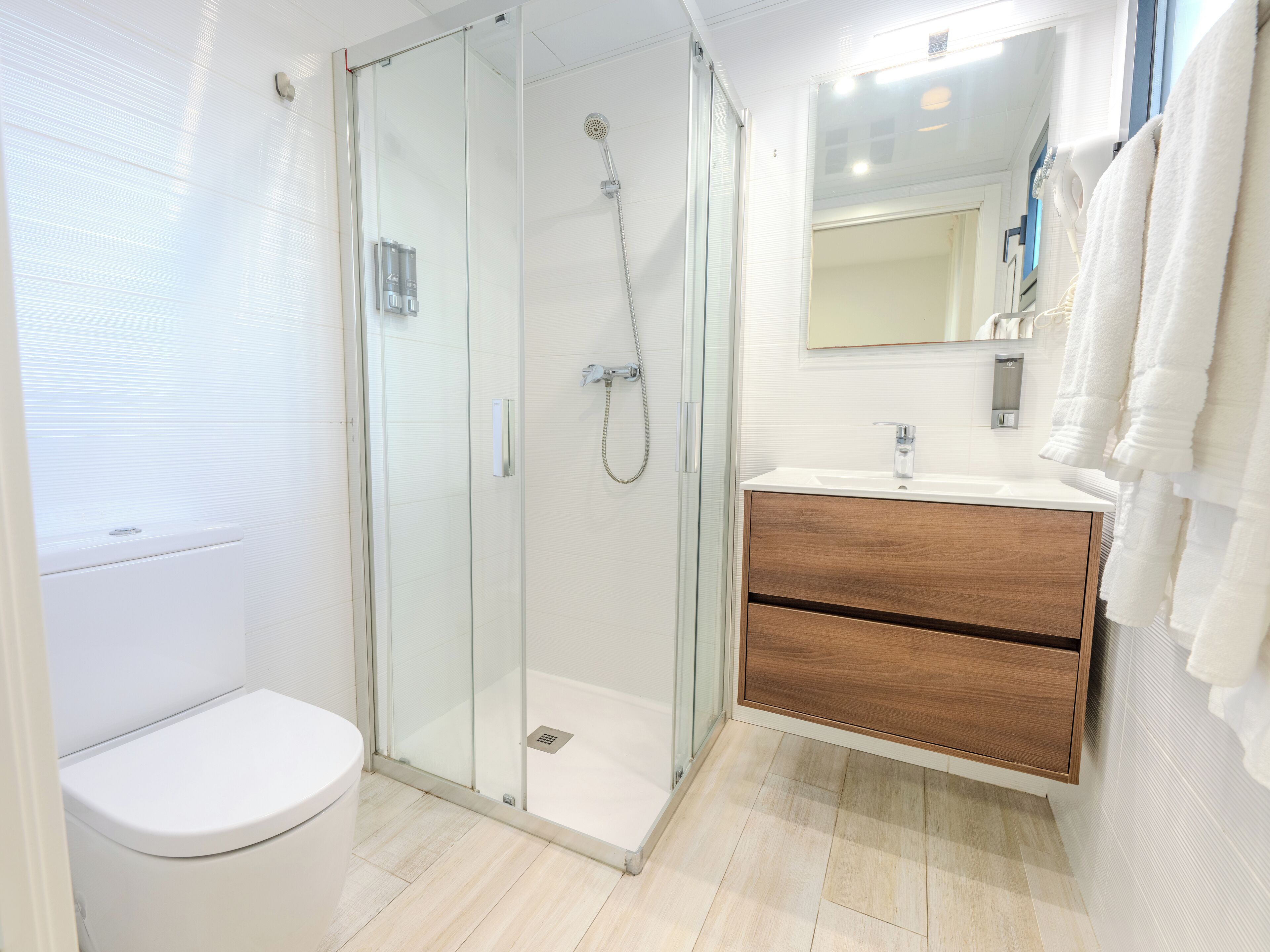 Un baño moderno con ducha envuelta en vidrio, inodoro blanco, tocador de madera y espejo sobre el lavamanos.