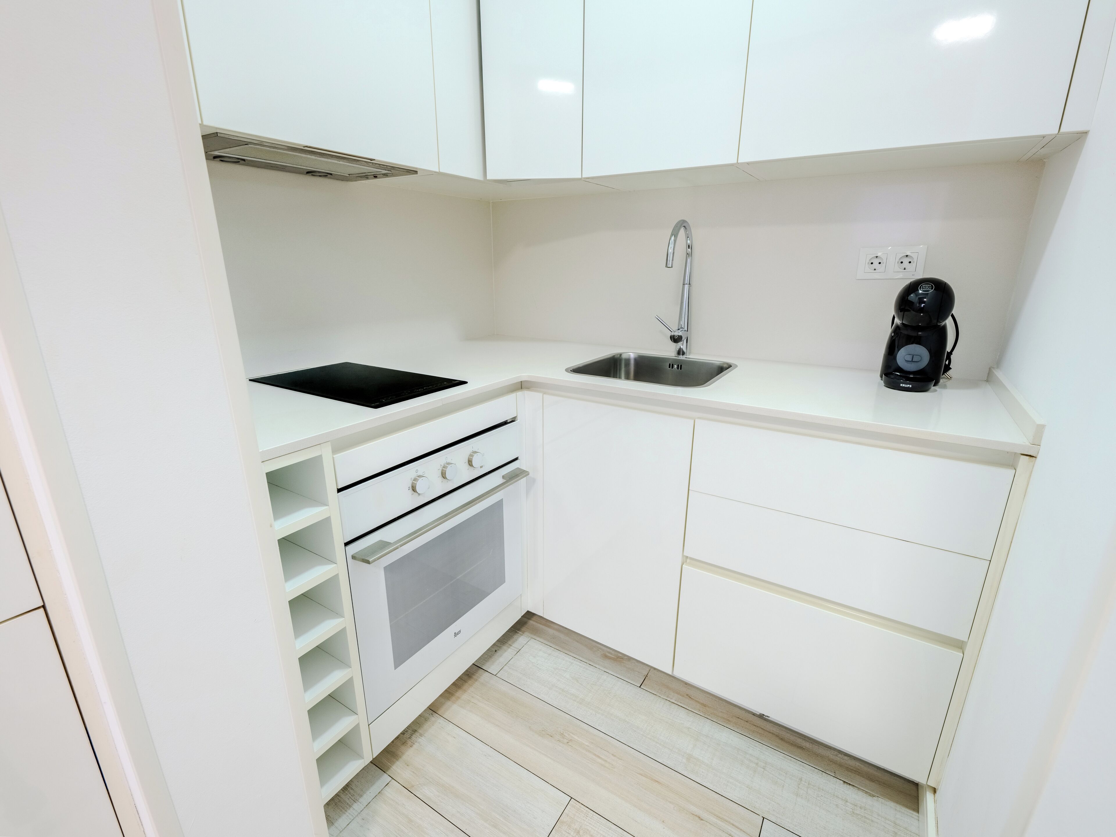 A compact kitchen with white cabinets, a black induction cooktop, a stainless steel sink, and a built-in oven.