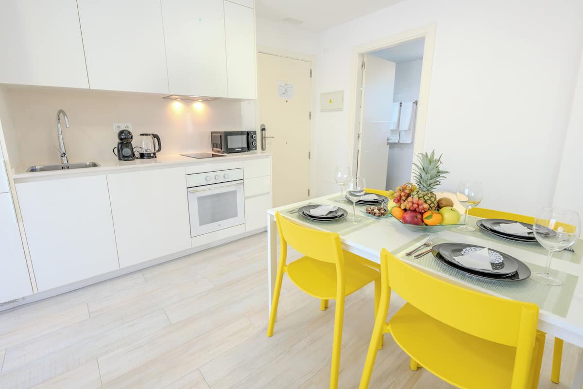A modern kitchen with white cabinets, a microwave, and a sink. There is a dining table with yellow chairs and a fruit bowl.