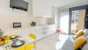 Een moderne keuken met witte kasten, een magnetron en een waterkoker. Er is een eettafel met gele stoelen en een grijze bank met gele kussens.