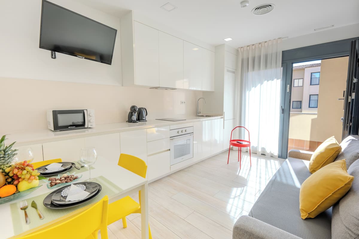 A modern kitchen with white cabinets, a microwave, and a kettle. There is a dining table with yellow chairs and a grey sofa with yellow cushions.