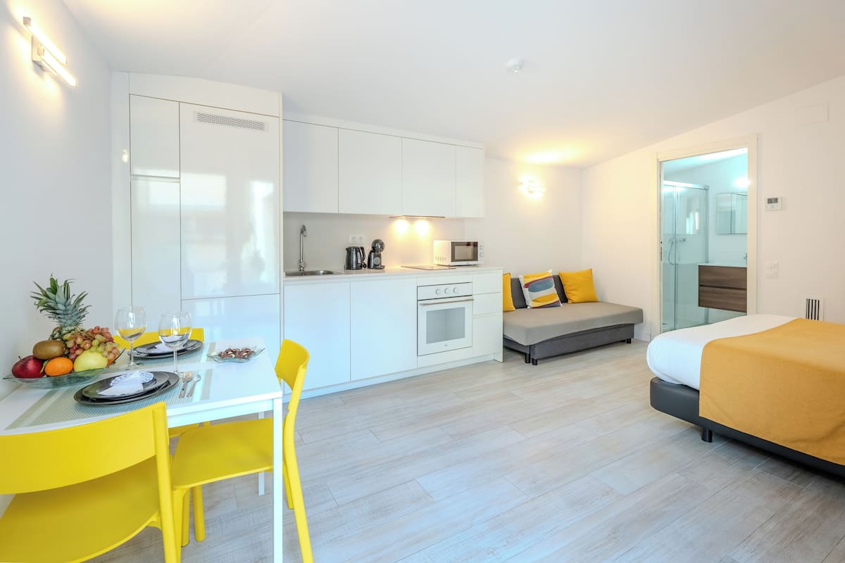A modern hotel room with a kitchenette, a dining area with yellow chairs, a bed with a yellow blanket, and a glass shower.